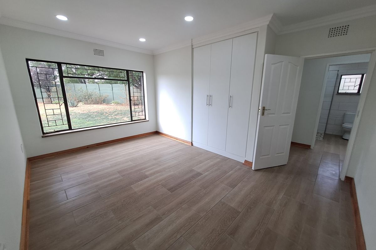 A bright, spacious bedroom featuring modern tiled flooring, built-in cupboards, recessed downlights, and large windows with security bars, leading to an en-suite bathroom.