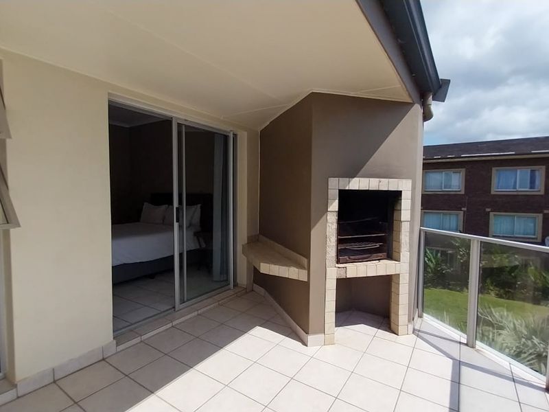 A covered balcony featuring tiled flooring, a built-in braai, and a glass balustrade, providing an outdoor space accessible via sliding doors.