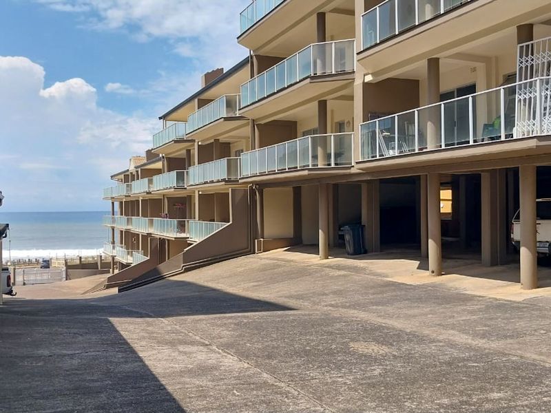 Exterior view of the apartment complex featuring tiered balconies with glass balustrades, concrete driveway, and covered parking area with ocean views.