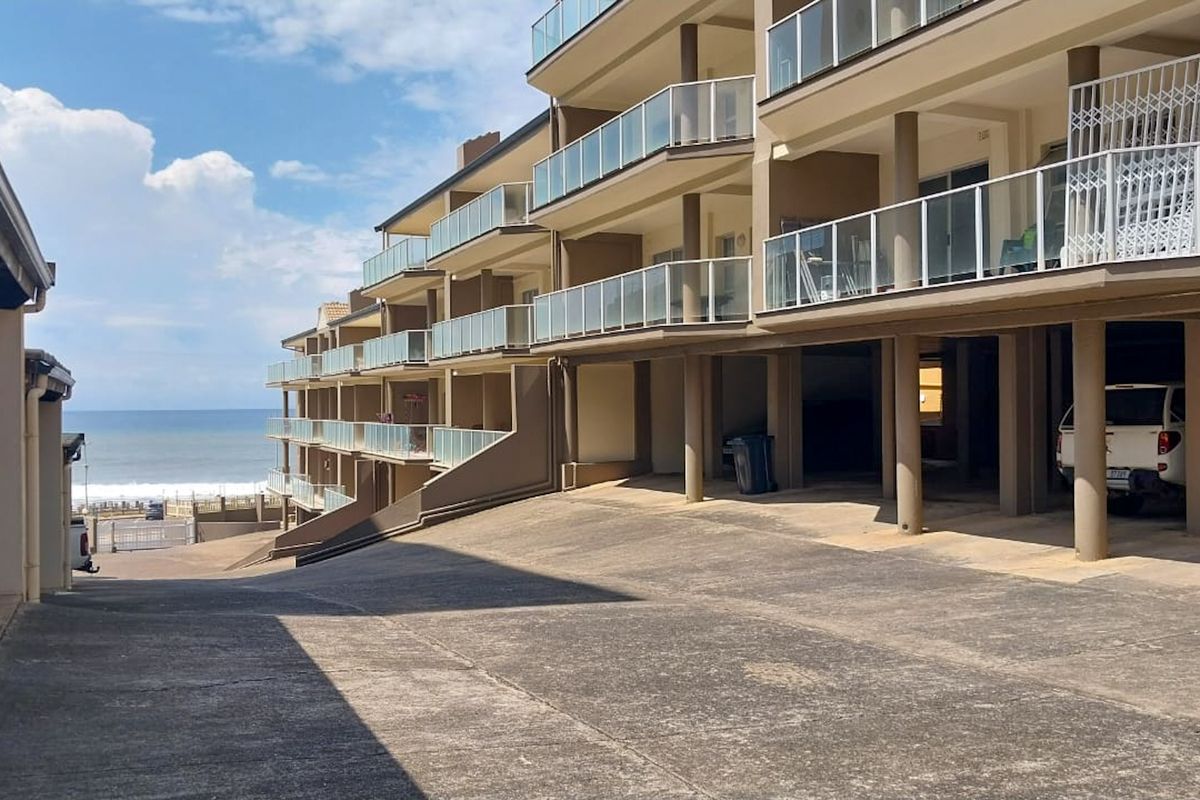 Exterior view of the apartment complex featuring tiered balconies with glass balustrades, concrete driveway, and covered parking area with ocean views.