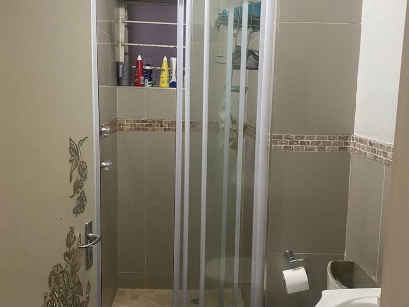 A compact bathroom featuring a shower enclosure, pedestal basin, and toilet with neutral wall tiling and a small window for ventilation.