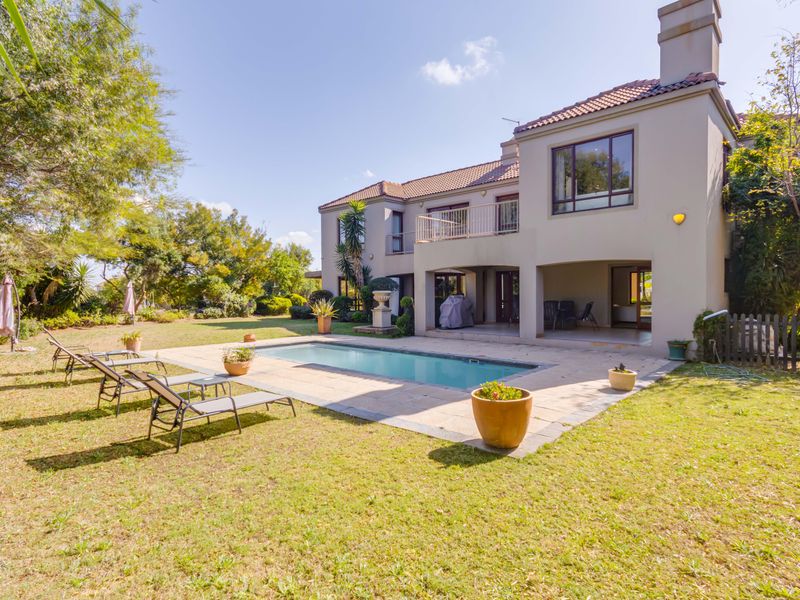 Rear view of a two-story house featuring a swimming pool, paved pool deck, covered patio, and a balcony overlooking the landscaped garden.