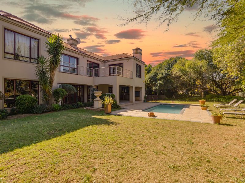 Rear view of a two-story house featuring a swimming pool, paved patio area, and a spacious landscaped garden at sunset.