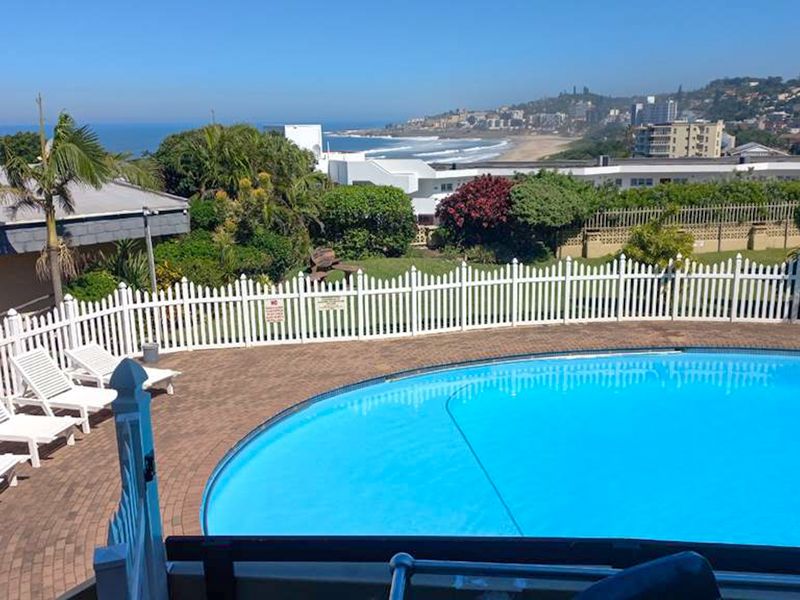 A well-maintained swimming pool area featuring a paved deck, white picket fencing, and scenic coastal views in the background.