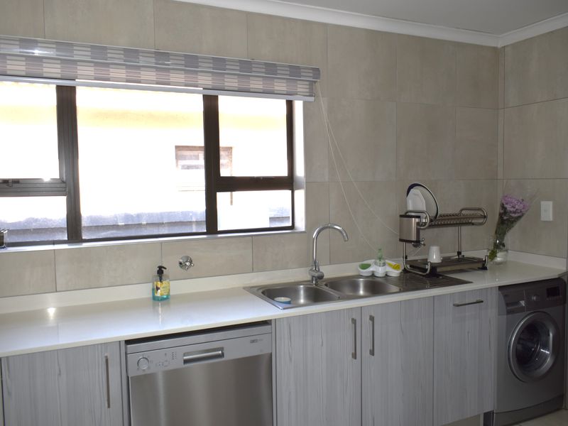 A modern kitchen area featuring light wood-grain cabinetry, stone countertops, a double sink, and integrated laundry and dishwashing appliances.