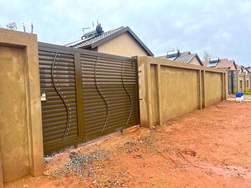Front view of the property featuring a secure metal sliding gate, high perimeter wall, and solar water heating system installed on the roof.