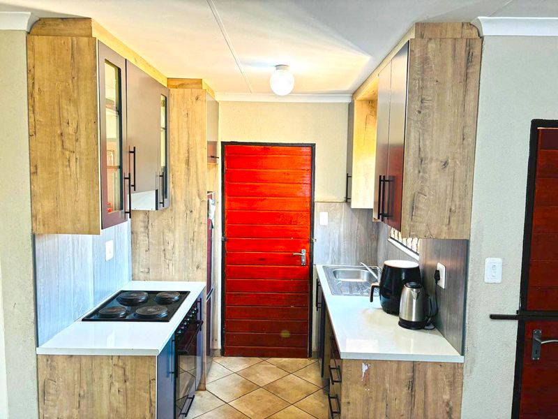Compact galley-style kitchen featuring wood-look cabinetry, integrated electric hob and oven, stainless steel sink, and tiled flooring.