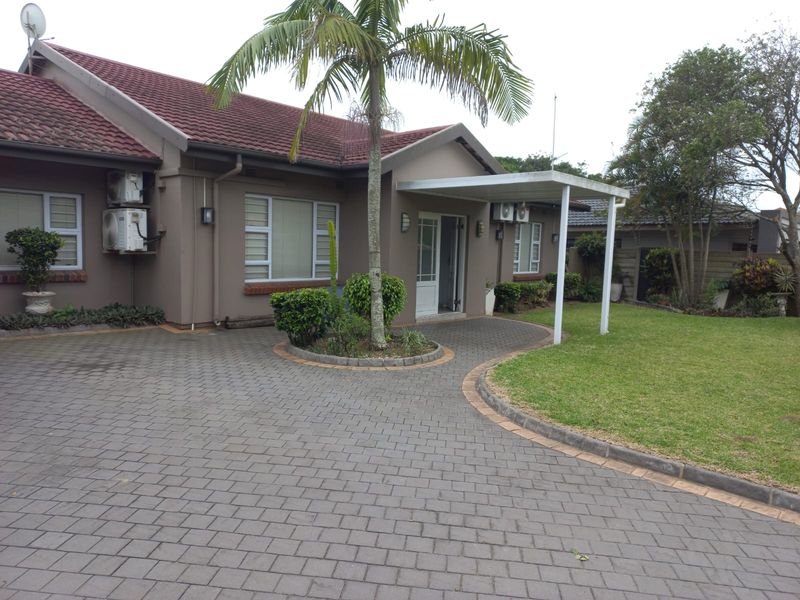A well-maintained single-story house featuring a paved driveway, covered entrance patio, tiled roof, and a landscaped garden area.