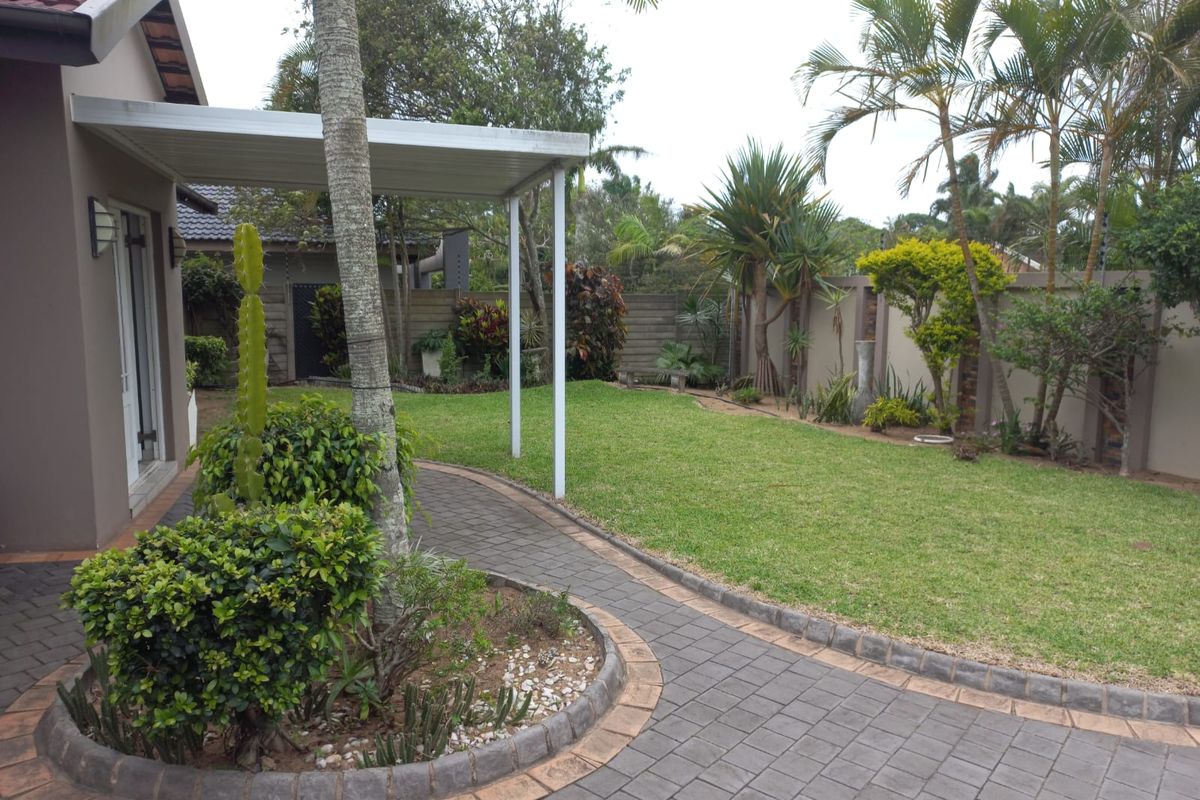A well-maintained rear garden featuring a paved walkway, manicured lawn, established landscaping, and a covered patio area.