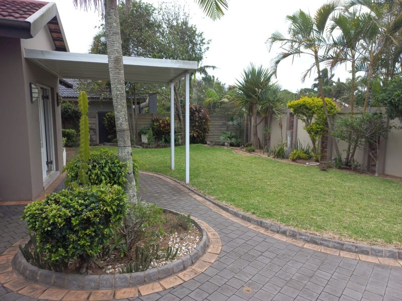 A well-maintained rear garden featuring a paved walkway, manicured lawn, established landscaping, and a covered patio area.