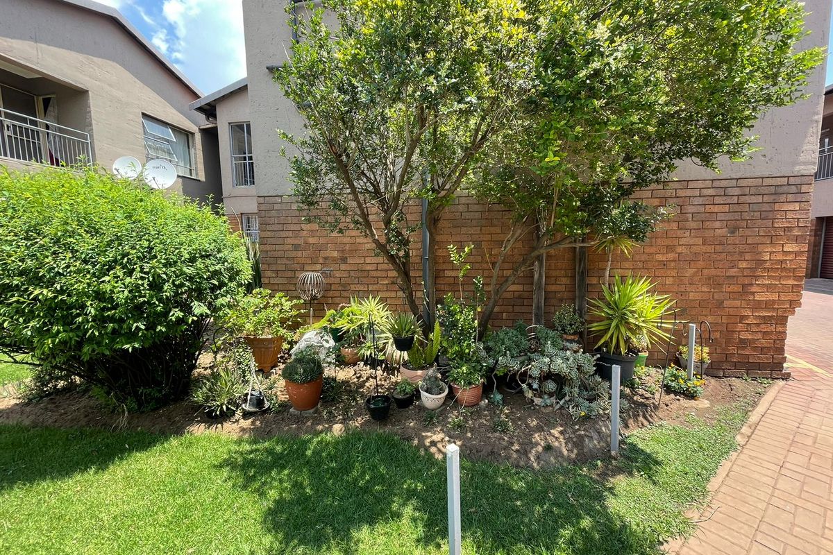 A view of the apartment building's rear exterior, featuring a brick facade, a small garden area with established trees, and a paved walkway.