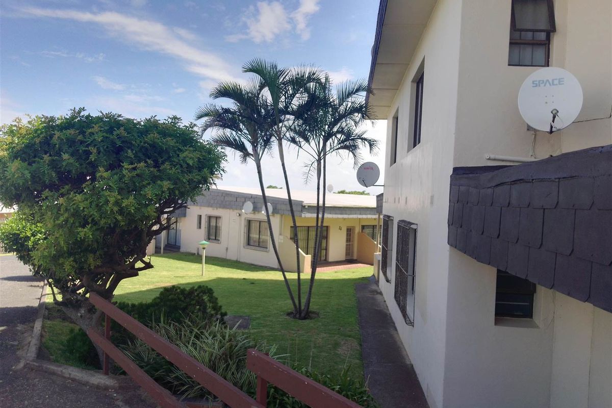 Exterior view of the apartment complex showing the rear garden area, paved walkways, and building facade with security bars and satellite dishes.