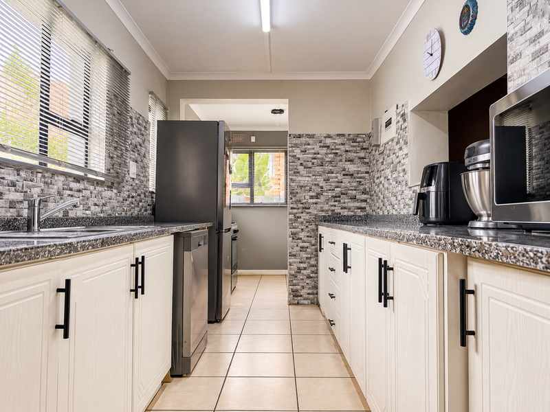 A functional galley-style kitchen featuring granite countertops, tiled backsplash, ample cabinetry, and tiled flooring with natural light from large windows.
