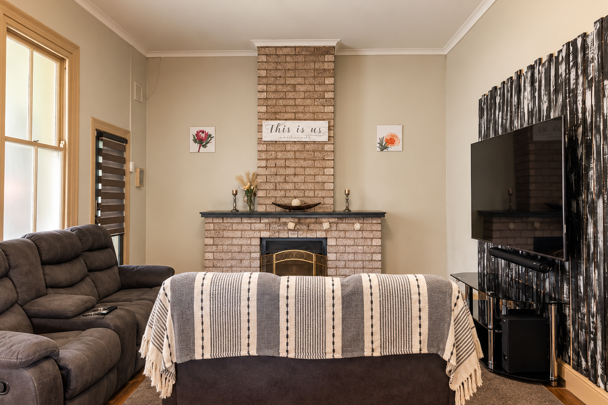 A cozy living room featuring a prominent brick fireplace, timber window frames, and warm timber flooring, creating a comfortable and inviting atmosphere.