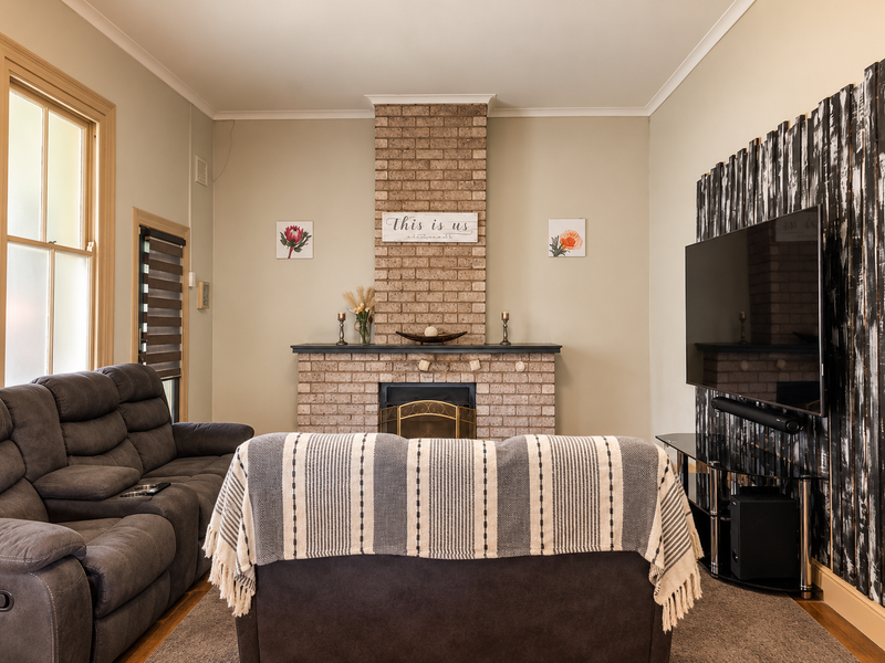 A cozy living room featuring a prominent brick fireplace, timber window frames, and warm timber flooring, creating a comfortable and inviting atmosphere.