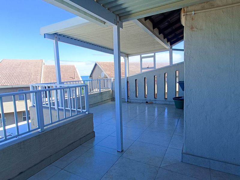 A spacious covered balcony featuring tiled flooring, metal railings, and a corrugated roof, offering an open outdoor area with coastal views.