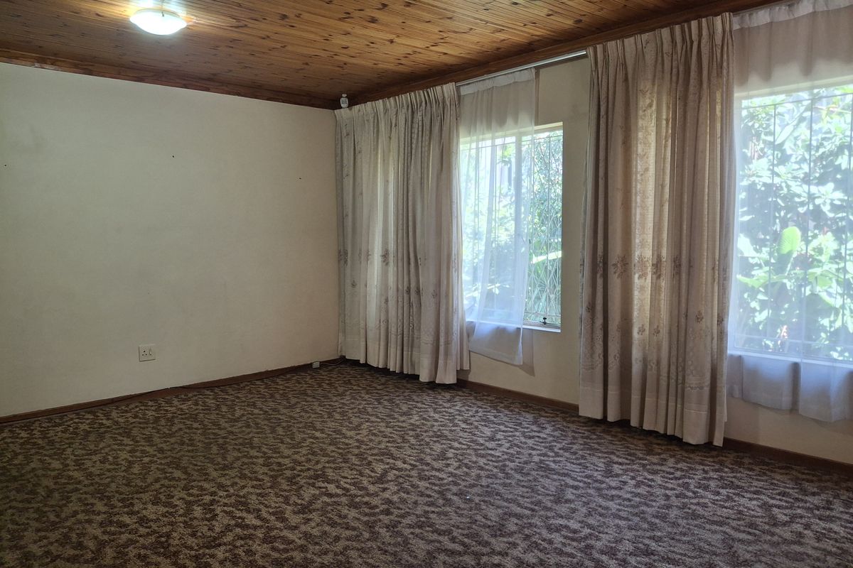 A spacious, empty living room featuring a timber-clad ceiling, patterned wall-to-wall carpeting, and large windows with integrated security bars.