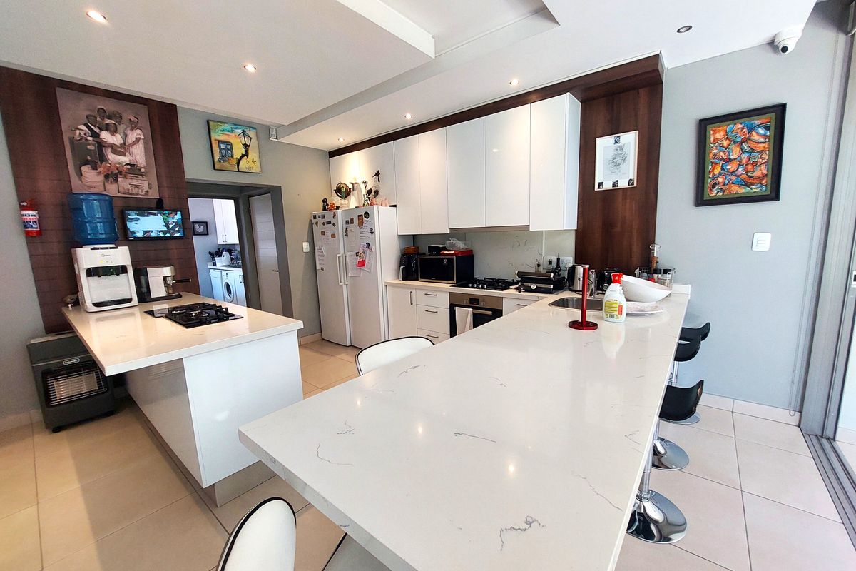 Modern kitchen featuring a large stone-topped island, sleek white cabinetry, built-in oven, gas hob, and recessed ceiling lighting with tiled flooring throughout.