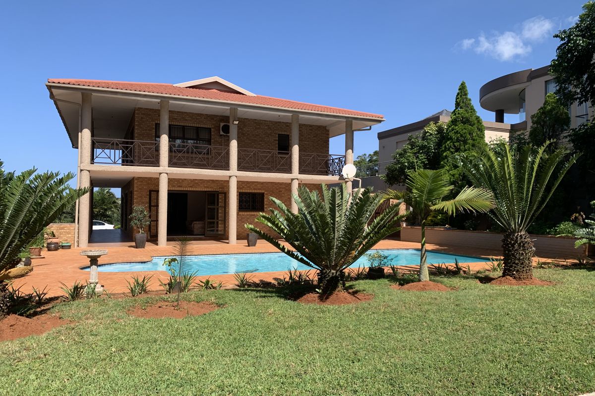 A spacious two-story brick house featuring a large swimming pool, covered patio areas on both levels, and a well-maintained garden.