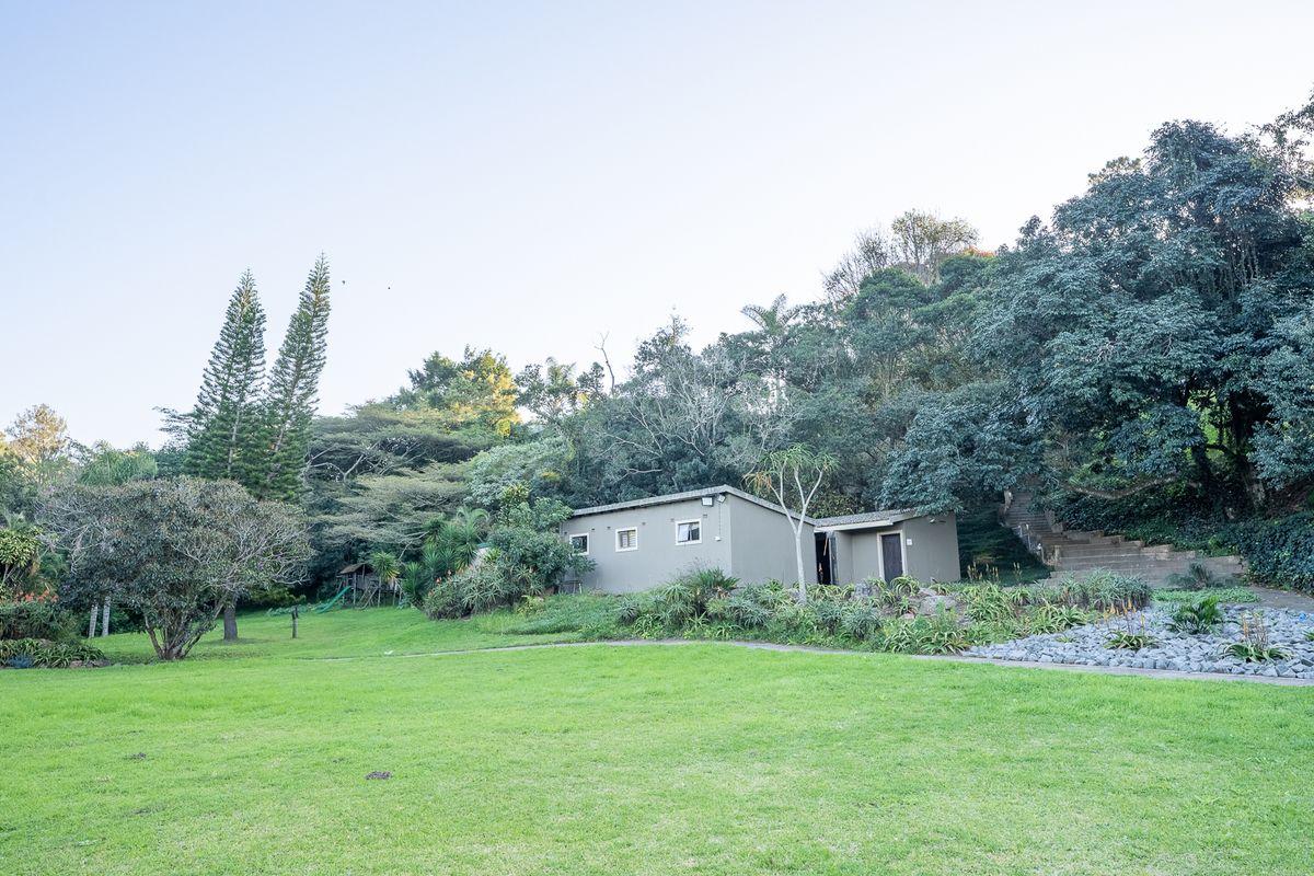 A spacious rear garden featuring a well-maintained lawn, mature trees, stone landscaping, and concrete stairs leading up the hillside.