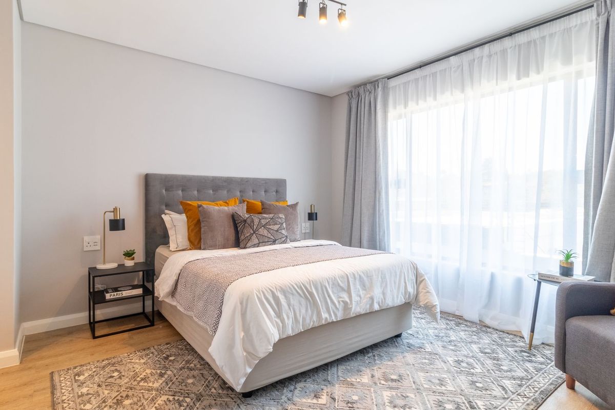 A bright and modern bedroom featuring light wood-look flooring, neutral wall tones, and large windows with sheer curtains allowing for ample natural light.