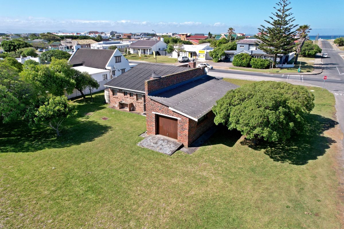 Aerial view of a brick house with a corrugated metal roof, attached garage, and a spacious surrounding garden in a coastal neighborhood.