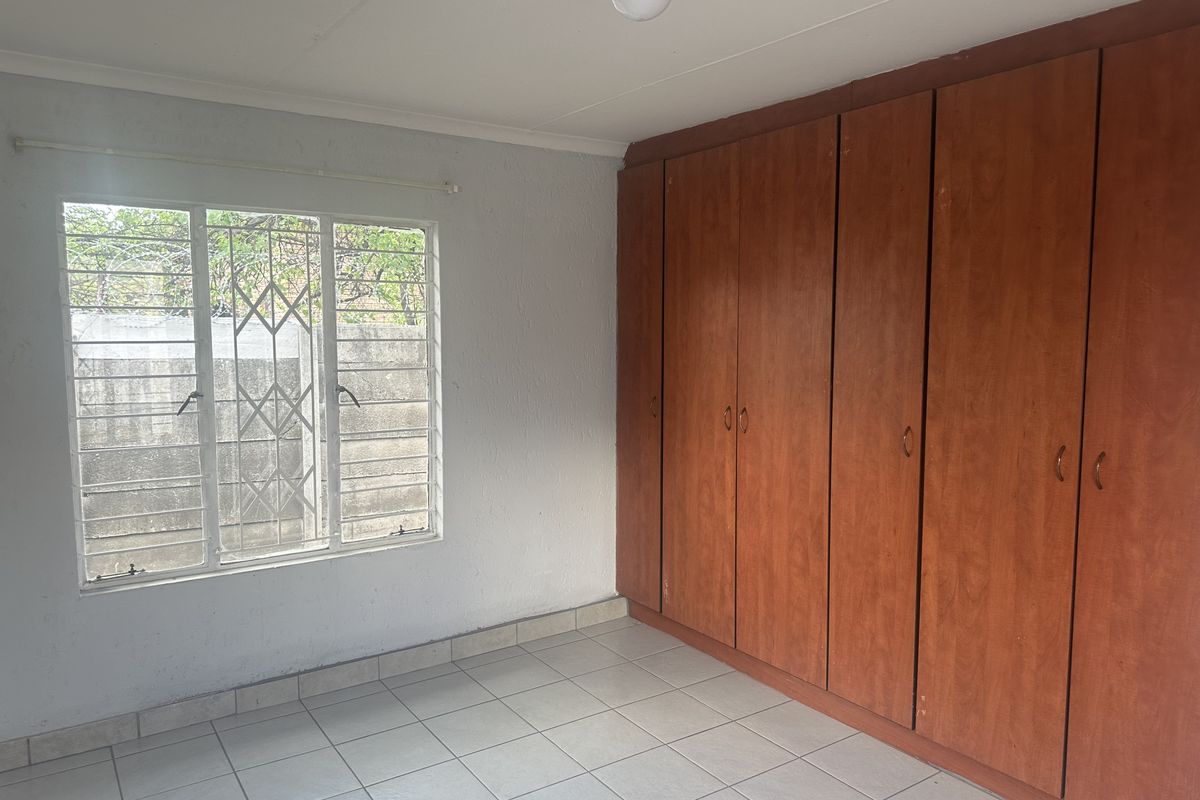 An empty bedroom featuring tiled flooring, built-in wooden cupboards, and a window fitted with security bars.