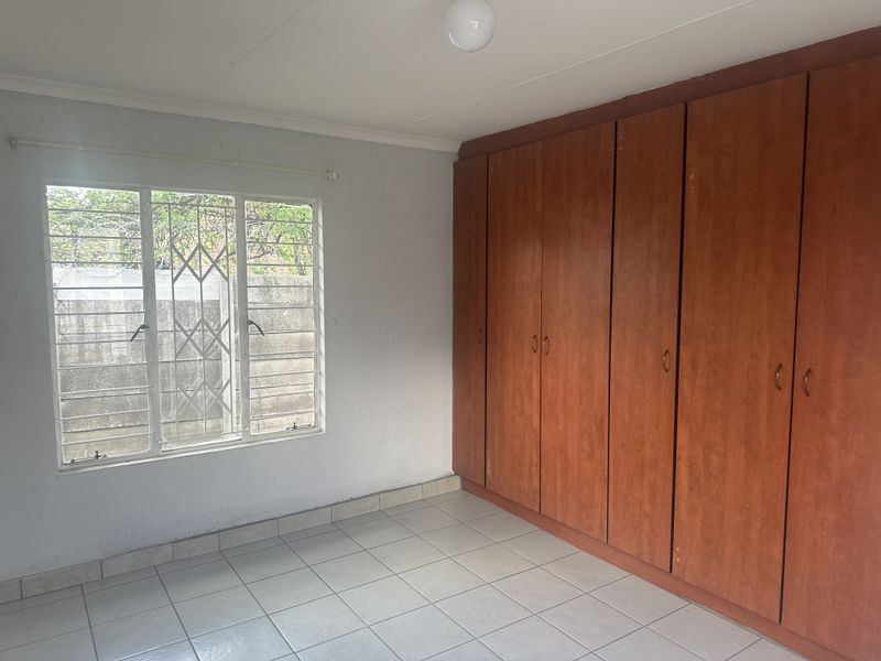 An empty bedroom featuring tiled flooring, built-in wooden cupboards, and a window fitted with security bars.