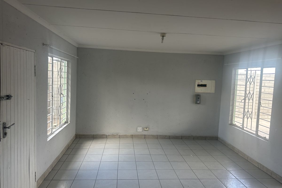An empty, neutrally painted living area featuring tiled flooring, security bars on windows, and a wall-mounted electrical distribution board.