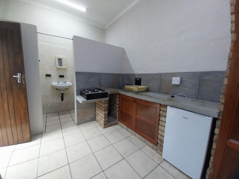 A compact kitchen area featuring a gas hob, concrete countertop, brick-based cabinetry, and an adjacent wall-mounted basin with tiled flooring throughout.