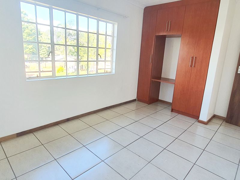 A bright, empty bedroom featuring tiled flooring, a large grid-pattern window, and built-in wooden cupboards.
