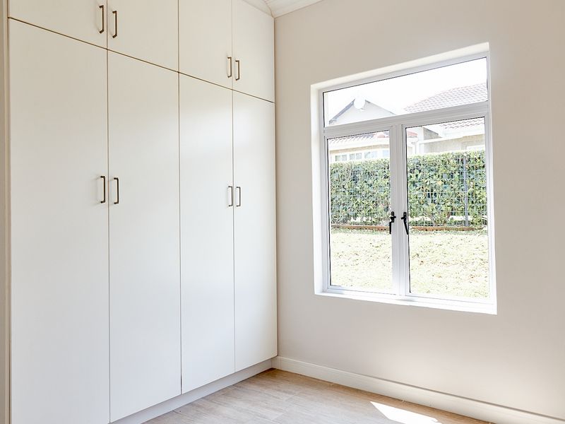 A bright, empty bedroom featuring modern white built-in cupboards, tiled flooring, and a large window overlooking the garden.