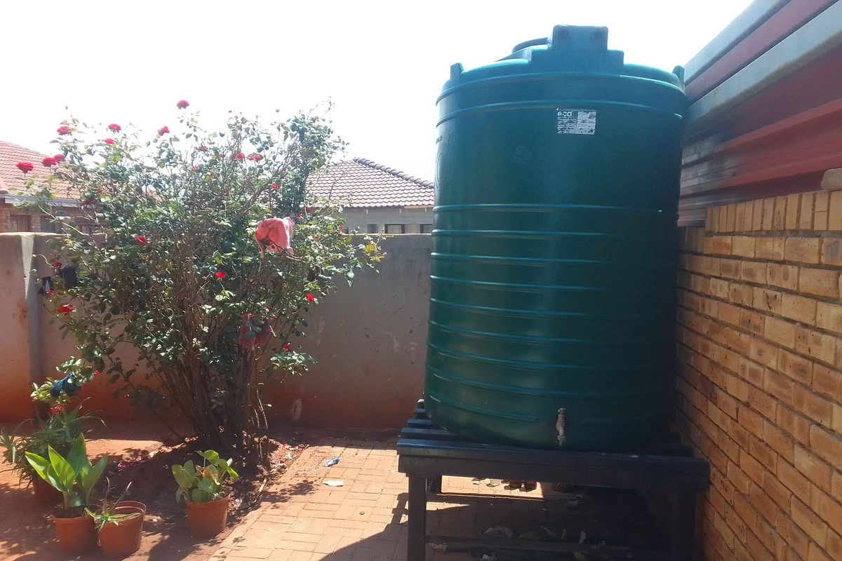 A rear exterior area featuring a large green water storage tank mounted on a metal stand against a brick boundary wall.