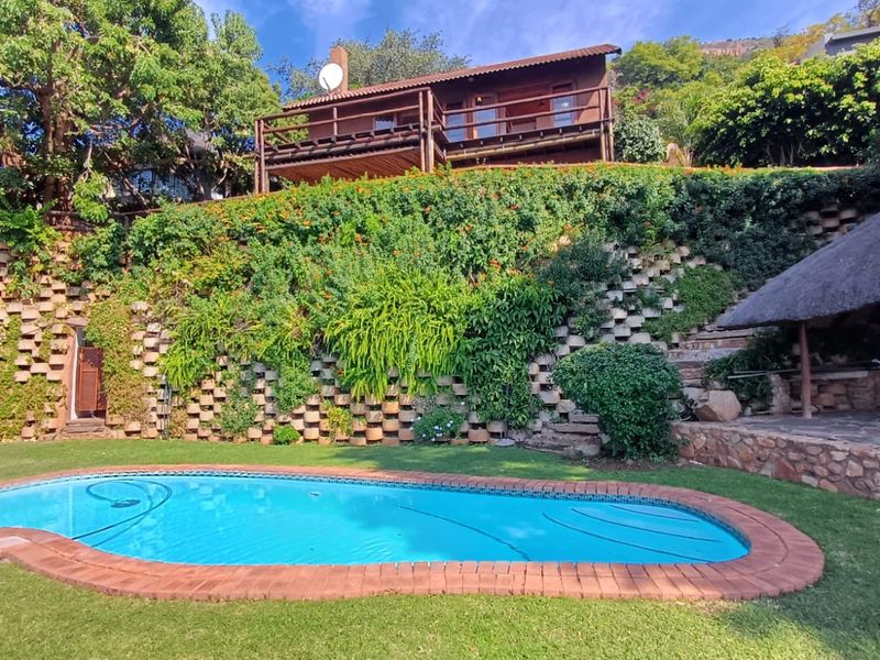 Outdoor pool area featuring a swimming pool with brick coping, a tiered retaining wall with lush greenery, and a thatched lapa structure.