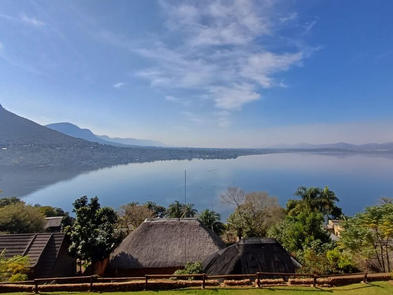 A scenic elevated view overlooking a large lake and surrounding mountains, featuring foreground thatched roof structures and a timber boundary fence.