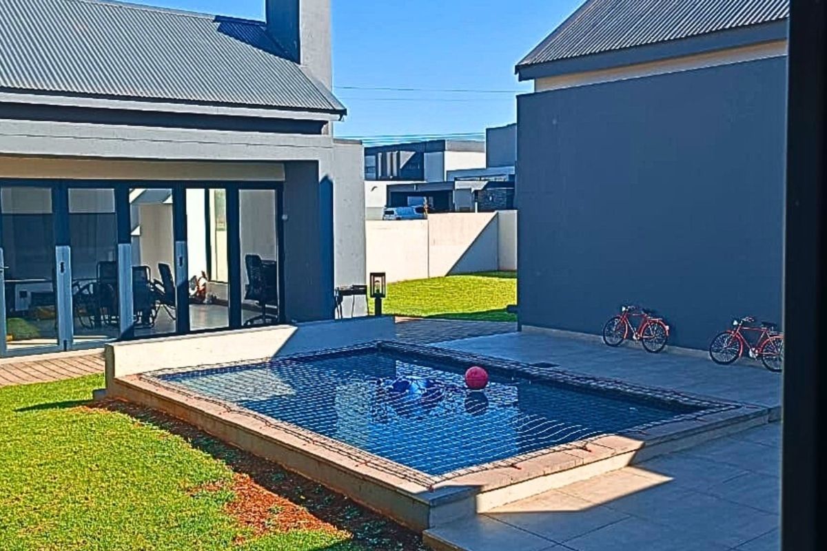 Rear exterior view featuring a swimming pool with a safety net, paved patio area, and a well-maintained lawn between two building structures.