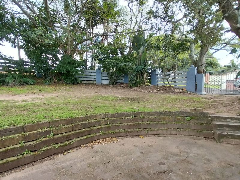 A rear garden area featuring a curved tiered retaining wall, concrete patio space, mature trees, and perimeter boundary fencing.