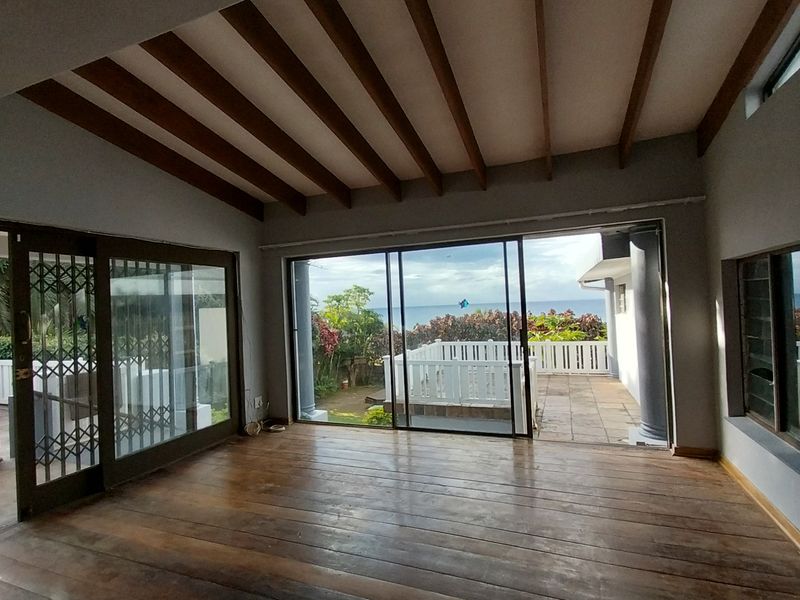 Spacious open-plan living area featuring exposed timber ceiling beams, hardwood flooring, and large sliding glass doors opening onto a patio with ocean views.
