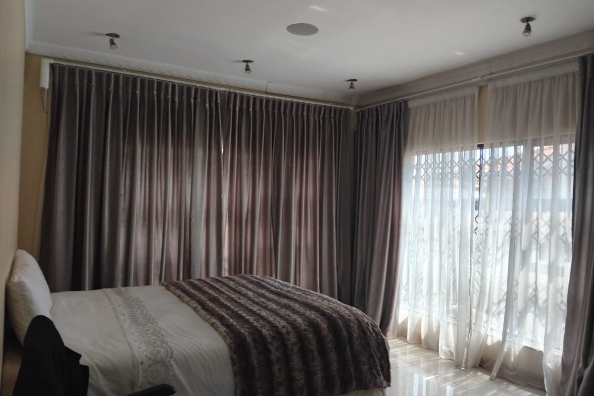 A well-lit bedroom featuring tiled flooring, recessed ceiling spotlights, and large windows fitted with security gates and full-length curtains.
