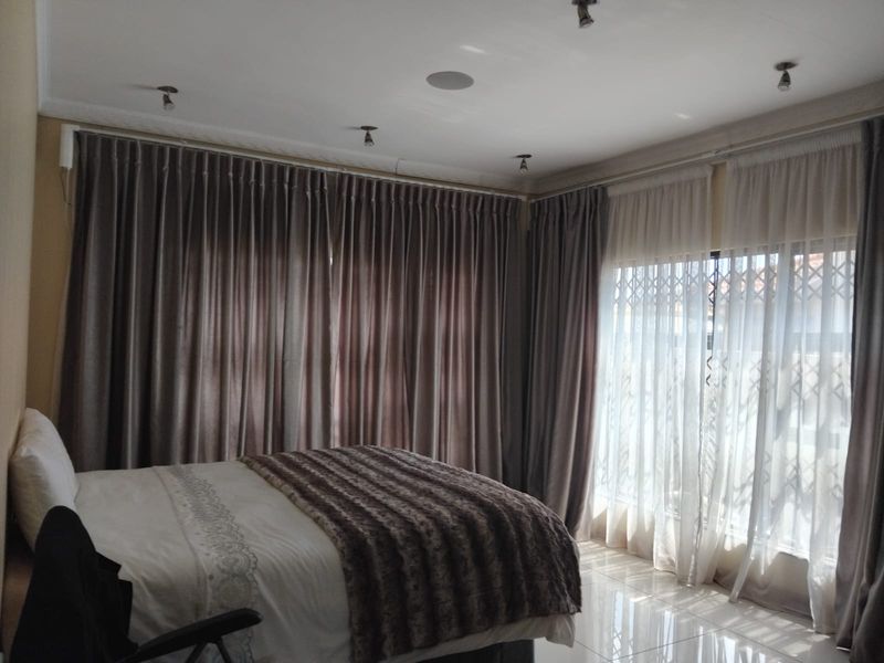 A well-lit bedroom featuring tiled flooring, recessed ceiling spotlights, and large windows fitted with security gates and full-length curtains.