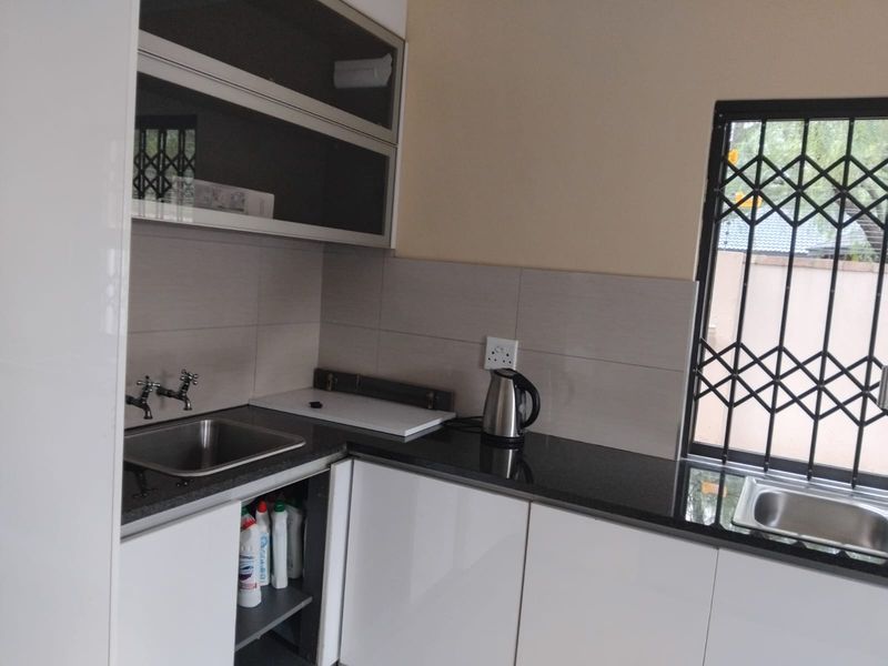 A functional scullery area featuring modern white cabinetry, dark granite countertops, dual stainless steel sinks, and a window with security bars.