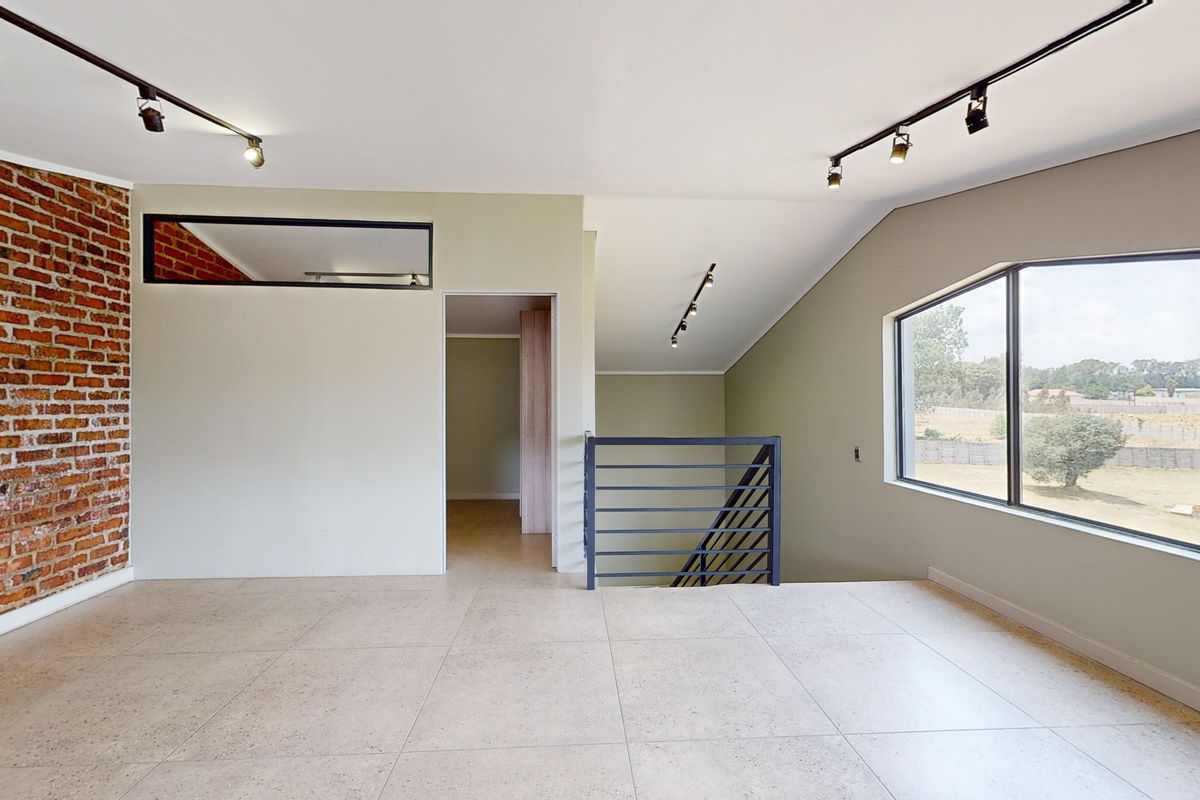 Spacious living area featuring an exposed brick feature wall, modern track lighting, large windows with scenic views, and a metal staircase railing.