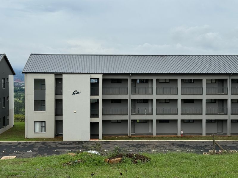 A modern multi-story apartment building featuring a grey metal roof, uniform balconies with metal railings, and an adjacent paved parking area.