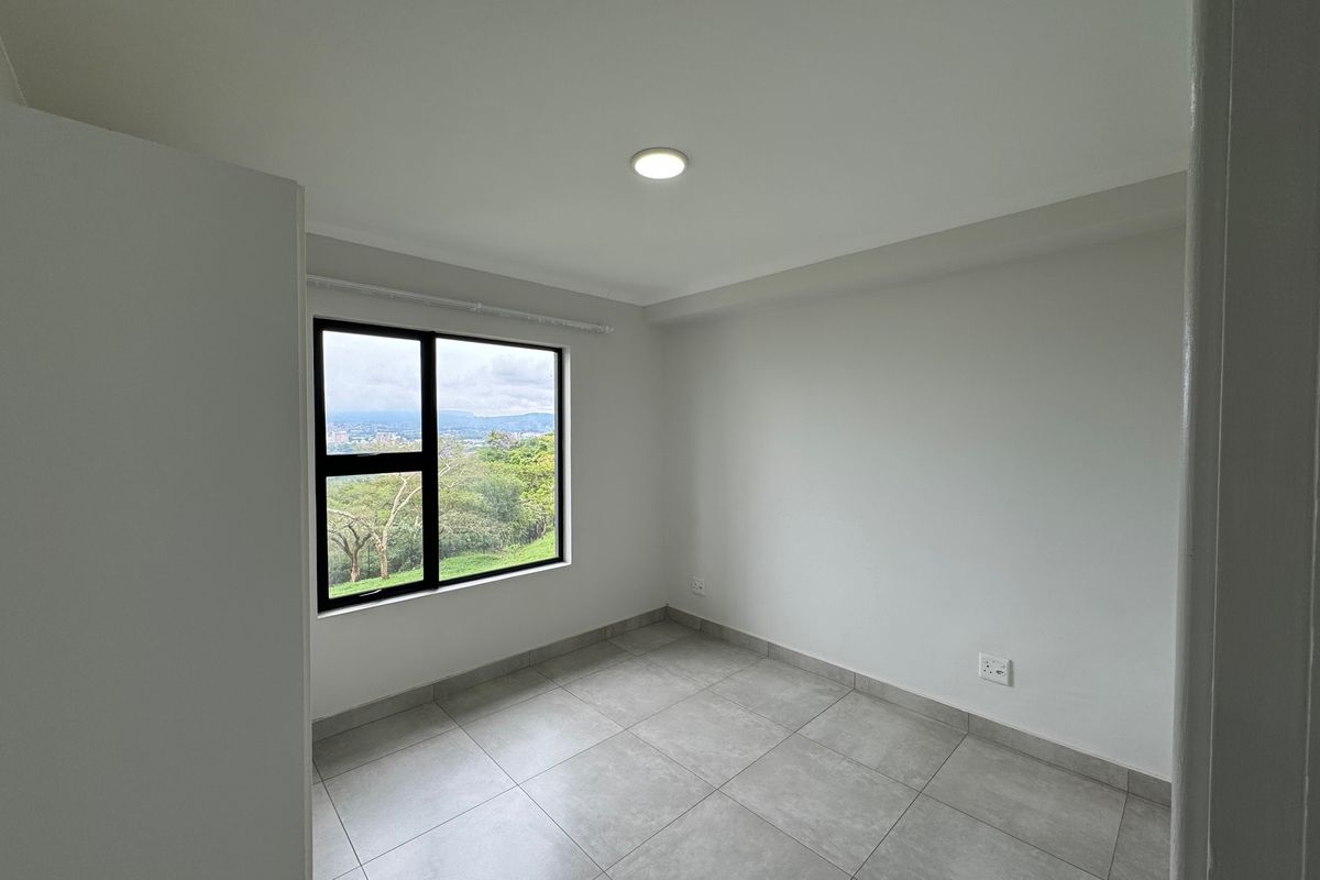 A bright, empty bedroom featuring neutral tiled flooring, a modern black aluminium window frame, and a scenic outdoor view.