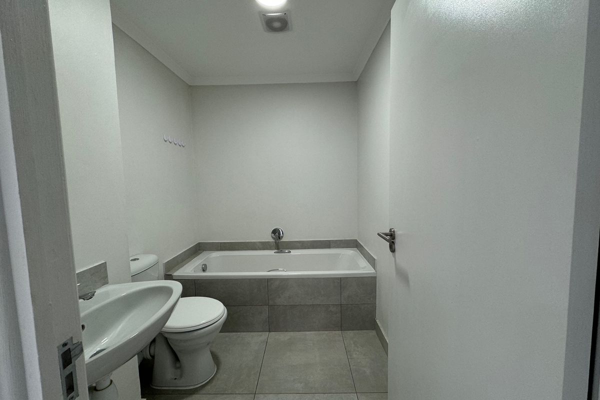 A clean, neutral bathroom featuring a built-in bathtub, pedestal washbasin, and tiled flooring, offering a functional and well-maintained space.