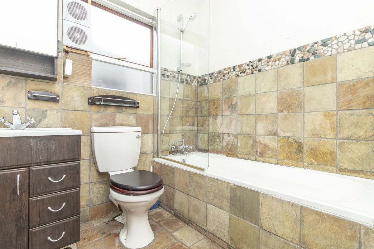 A dated bathroom featuring a combined shower and bathtub, vanity cabinet, and toilet, finished with rustic stone-style wall and floor tiles.