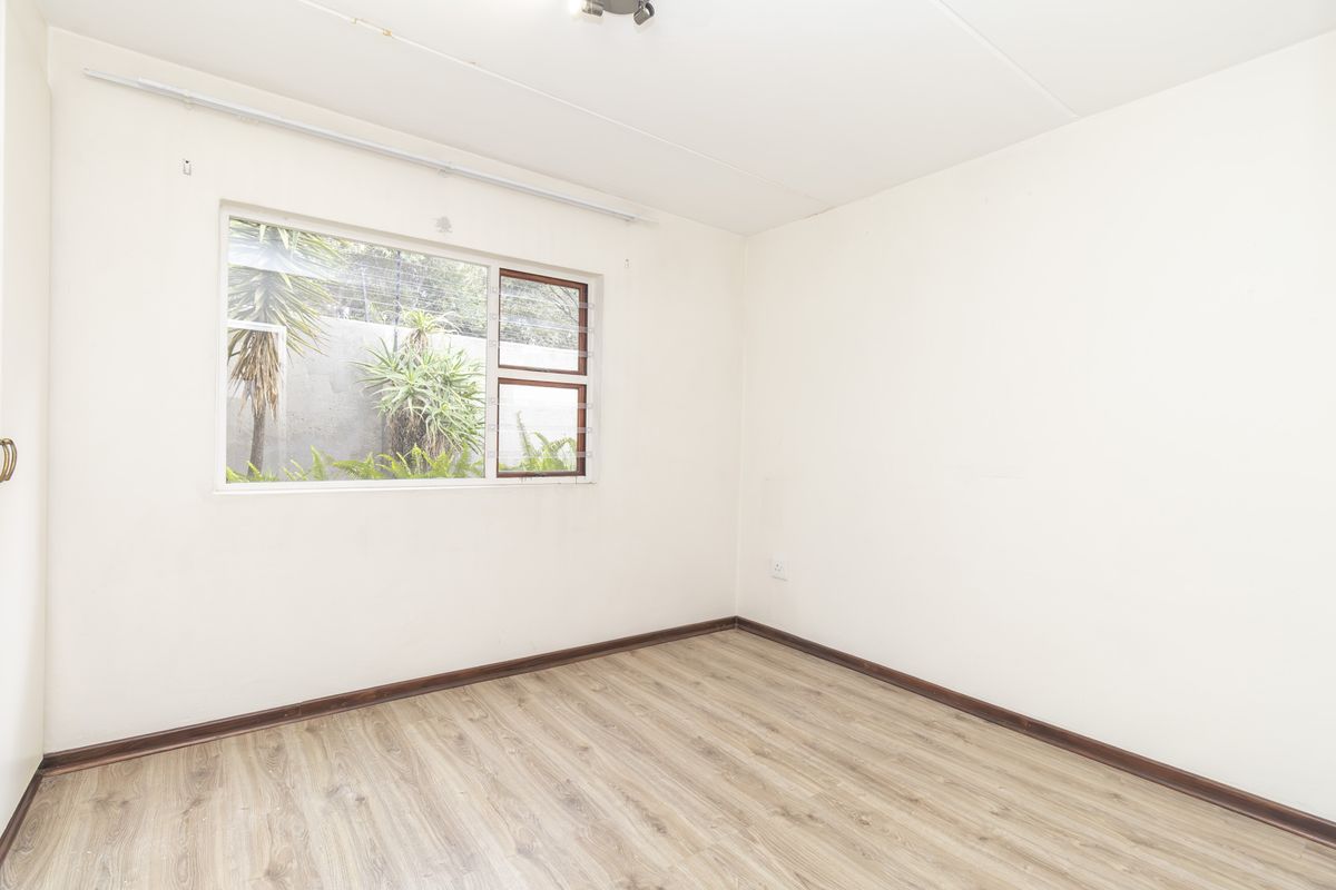 A bright, empty bedroom featuring wood-look laminate flooring, a large window with garden views, and neutral wall finishes.