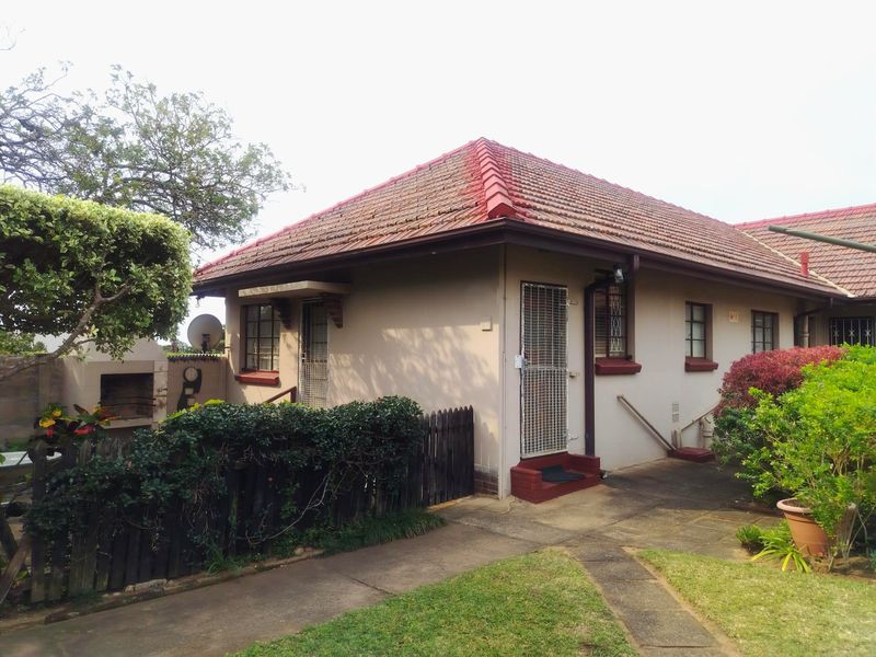 Rear exterior view of the apartment featuring a tiled roof, paved walkway, and a built-in braai area within a small garden setting.