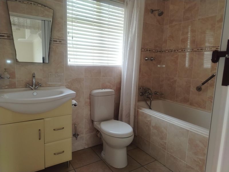 A functional bathroom featuring a vanity cabinet, ceramic basin, toilet, and a tiled bathtub area with a shower head and grab rail.