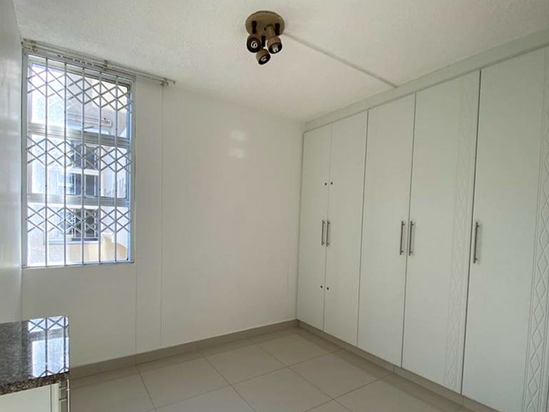 A bright, empty bedroom featuring neutral tiled flooring, ample built-in cupboard storage, and a window fitted with a security gate.